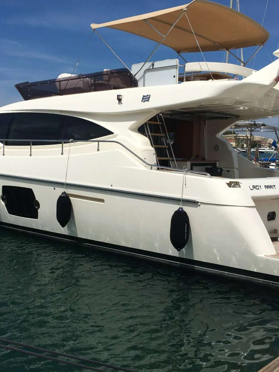 Een witte motorjacht met de naam "LADY MARIT" ligt aangemeerd in een haven, met een beige zonnescherm en zwarte stootwillen.
