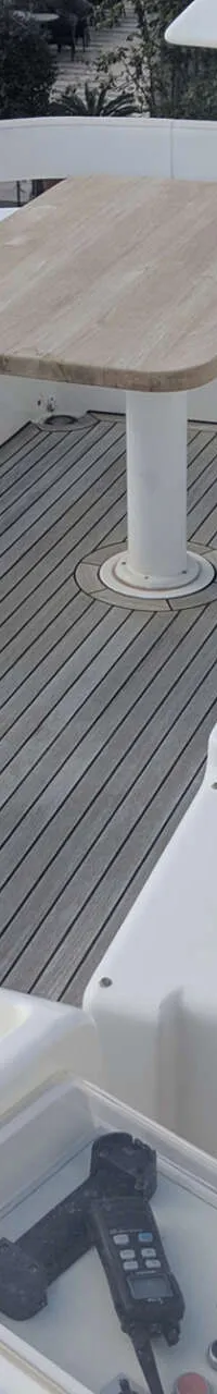 De foto toont een teakhouten dek met een houten tafel, witte banken, een trap en een stuurwiel.