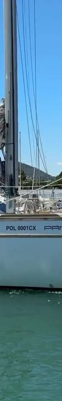 Een witte zeilboot met rode zeilen en de naam "PANGEA" vaart op helderblauw water, met bergen en een stad op de achtergrond.