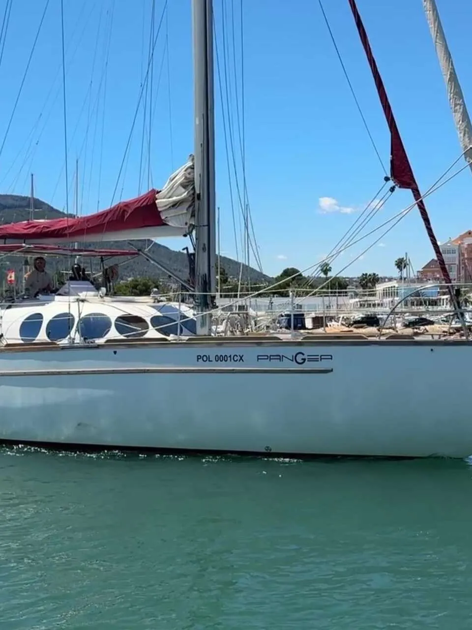 Een witte zeilboot met rode zeilen en de naam "PANGEA" vaart op helderblauw water, met bergen en een stad op de achtergrond.