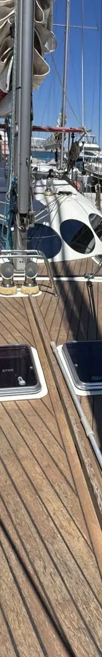 Een teak dek met twee luiken en een opgerold zeil op een mast, omringd door andere boten in een haven.