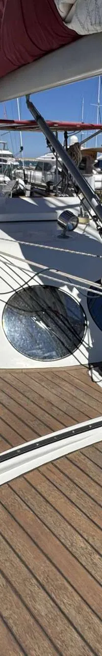Een dek van een zeilboot met een houten vloer, een witte opbouw met ronde ramen en een mast met touwen.