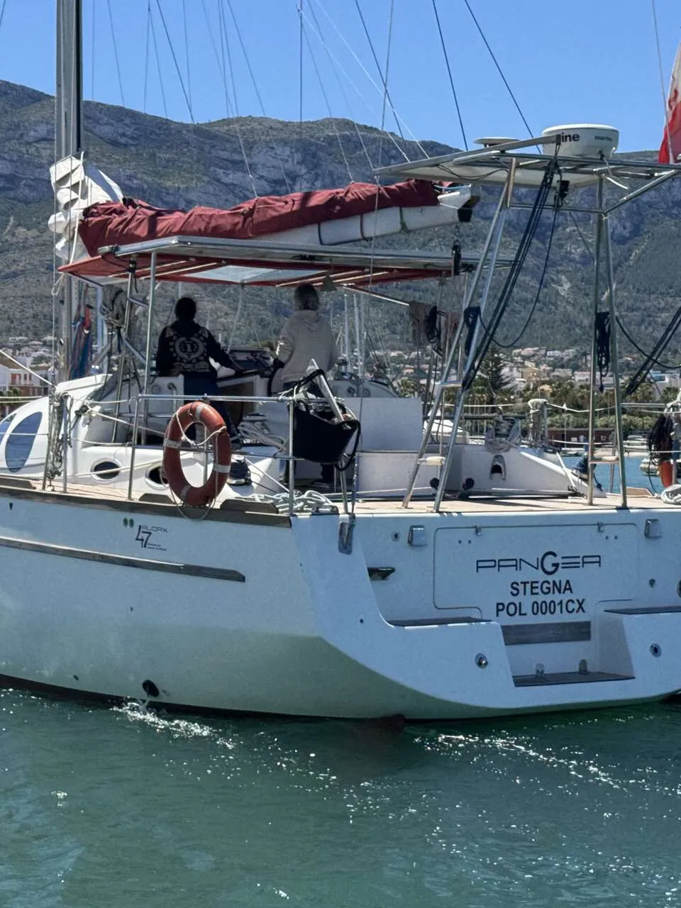 Een witte zeilboot met twee personen aan het roer vaart in een haven met bergen op de achtergrond. — Custom Built Audax 47
