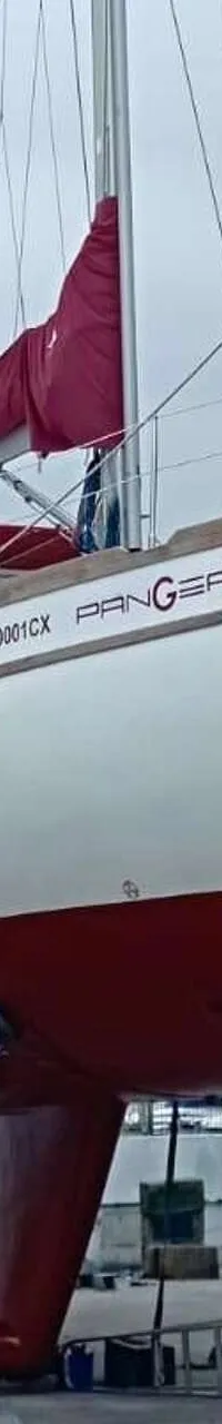 Een witte zeilboot met rode romp en kiel staat op bokken op de wal, met de naam "PANGEA" op de zijkant.
