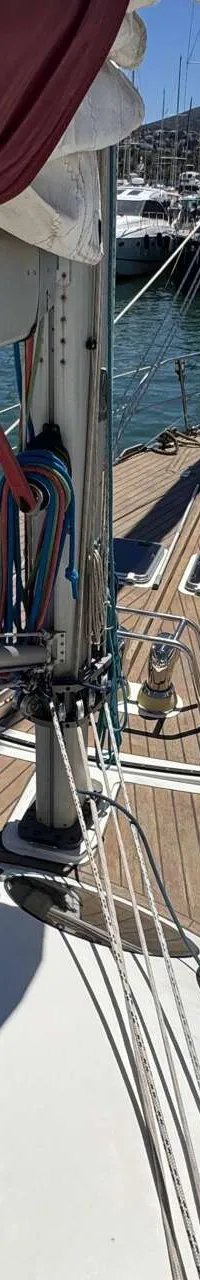 Een bovenaanzicht van het teakhouten dek van een zeilboot met een opgerold rood zeil, omringd door andere boten in een haven.