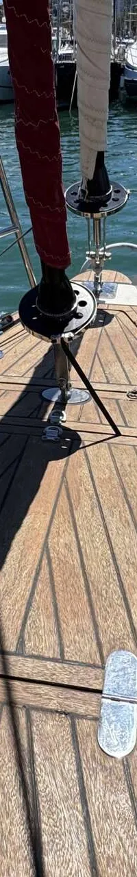 De foto toont het teakhouten voordek van een zeilboot met een rode en witte rolfok, reling en touwen, liggend in een haven.