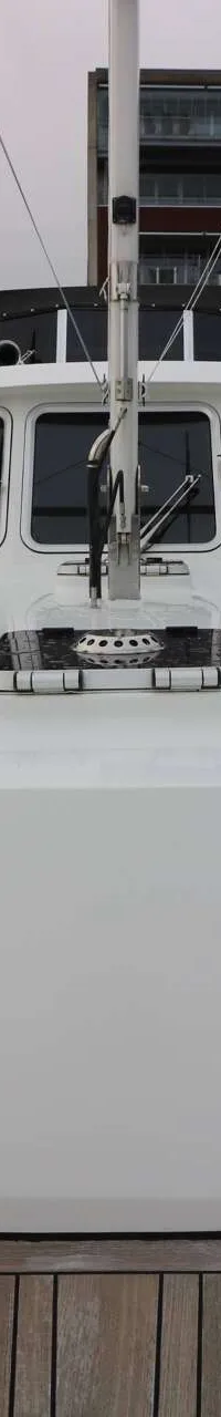 De foto toont de boeg van een witte motorboot met een houten dek, liggend in een haven met gebouwen op de achtergrond.
