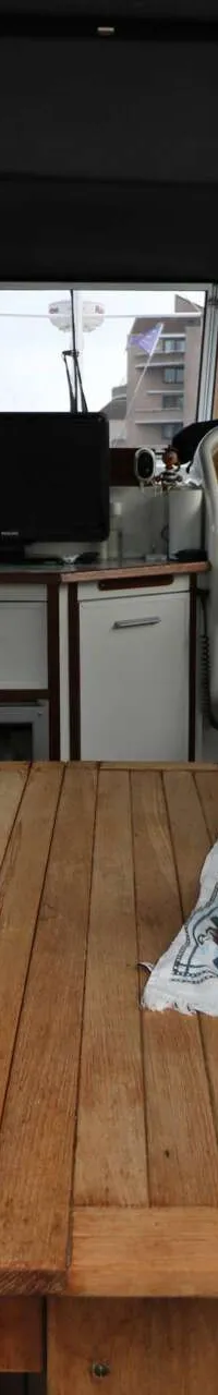 De foto toont het interieur van een boot met een houten tafel, stoelen met bladmotief kussens, een stuurstand en een monitor.