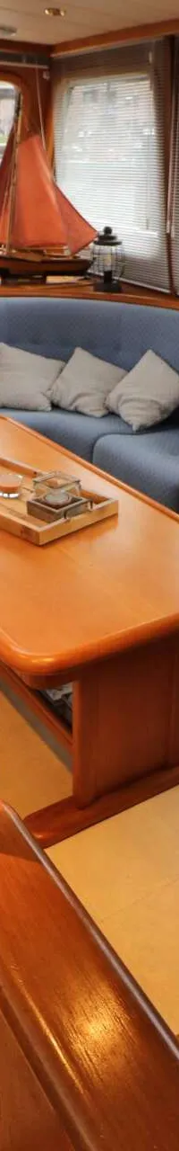 De foto toont het interieur van een boot met een stuurstand, een tafel, een fauteuil en twee banken met blauwe kussens.
