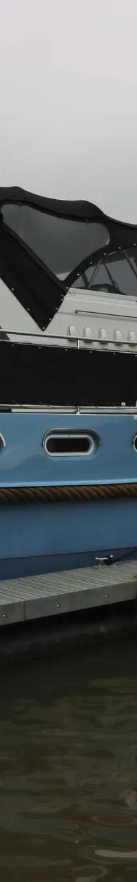 Een blauw-witte motorboot ligt afgemeerd aan een steiger, met een brug en gebouwen op de achtergrond.