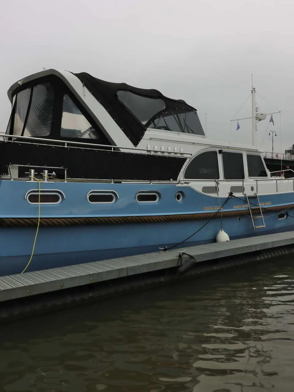 Een blauw-witte motorboot ligt afgemeerd aan een steiger, met een brug en gebouwen op de achtergrond.