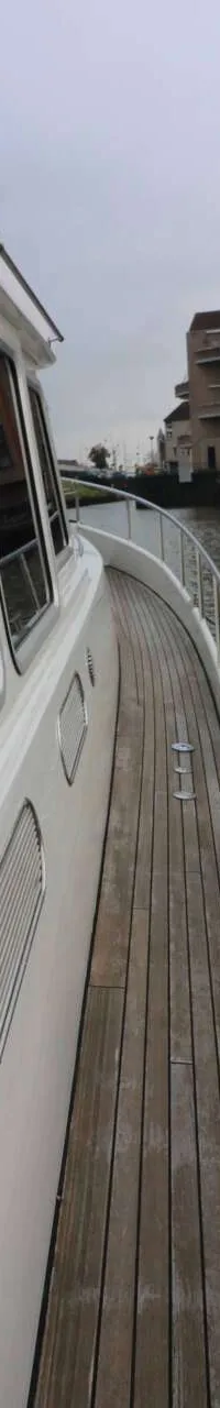 Een witte boot met houten dek ligt aangemeerd aan een steiger, met gebouwen en water op de achtergrond.