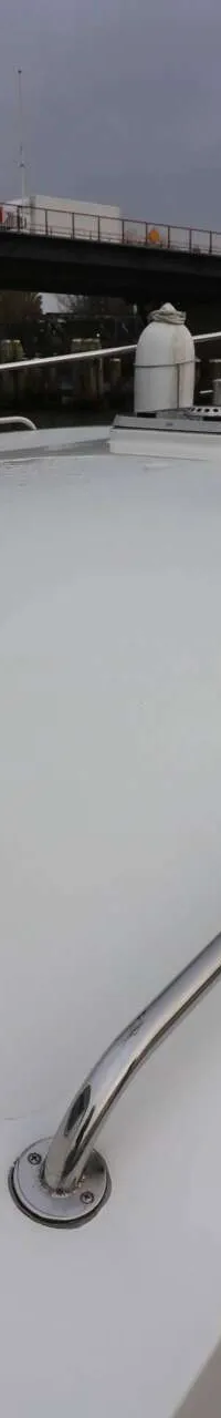 De foto toont het voordek van een boot met een wit dek, houten zijdekken en een reling, met een brug op de achtergrond.
