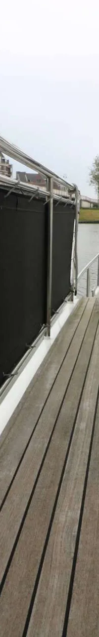 Een houten dek met zwarte zeilen aan de zijkanten en een reling, met een waterweg en gebouwen op de achtergrond.