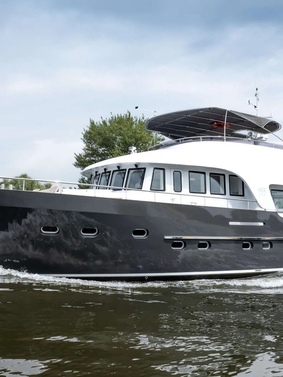 Een donkergrijze motorboot met een witte opbouw vaart over water, met een De Valk Yacht Brokers-logo rechtsboven.