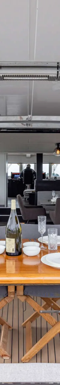 Een gedekte houten tafel met stoelen op het achterdek van een jacht, met uitzicht op water en begroeide oevers.