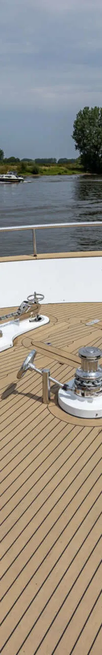 De boeg van een boot met een teakhouten dek, twee ankerlieren, een zitbank en een Nederlandse vlag, varend op een rivier.