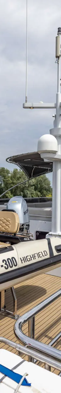 Een witte motorboot met een bijboot aan dek vaart op een rivier met bomen langs de oevers onder een bewolkte lucht.
