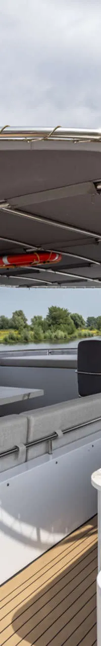De foto toont het achterdek van een jacht met een zithoek, tafel, stuurstand en een luifel, varend op een rivier.