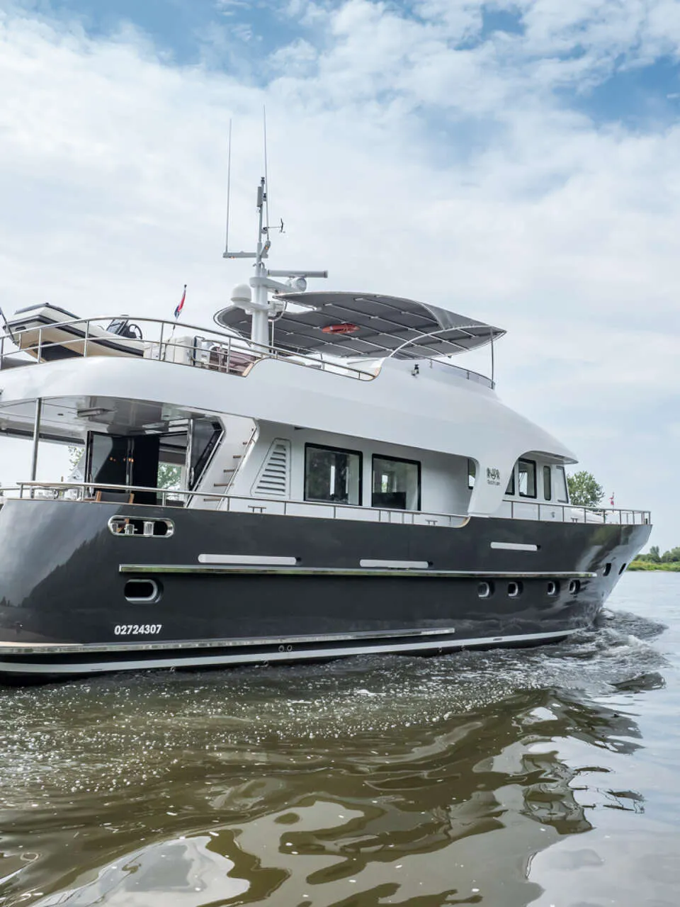 Een donkergrijze motorboot met witte opbouw vaart op een rivier onder een bewolkte hemel. — Bloemsma Trawler 70