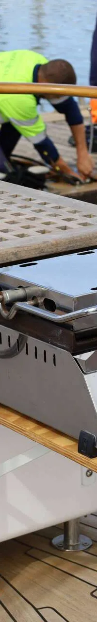 Een roestvrijstalen grill met twee rode knoppen is ingebouwd in een houten kast op een teakdek.