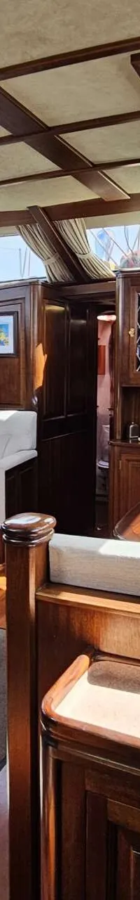 De foto toont het interieur van een boot met houten lambrisering, een zithoek met banken en een tafel, en ramen met…