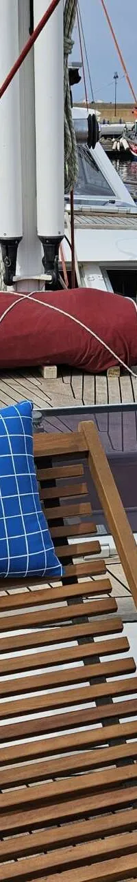 Het dek van een zeilboot met twee houten ligstoelen en blauwgeruite kussens, omringd door andere boten in een haven.