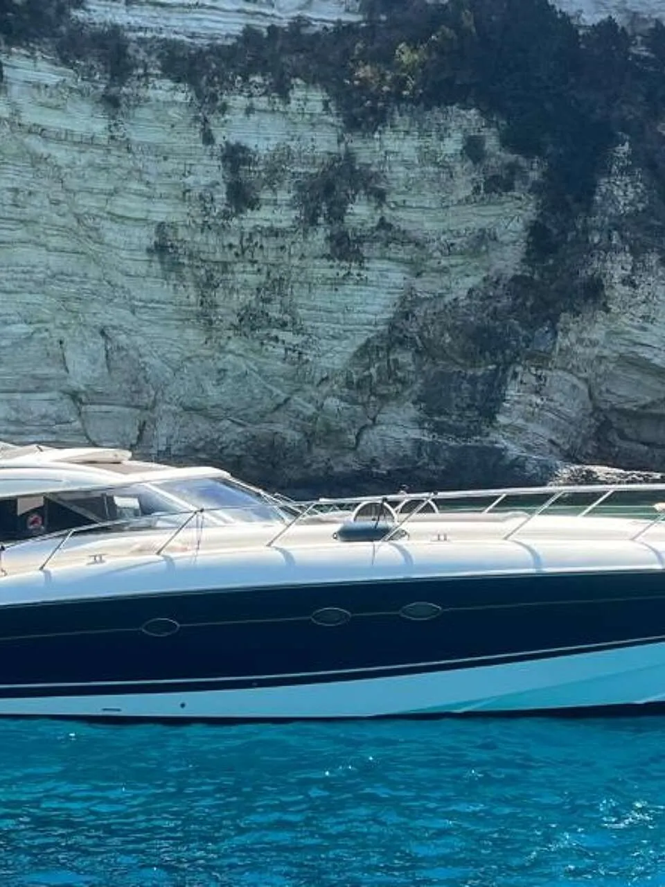 Een witte en donkerblauwe motorboot ligt in helderblauw water voor een rotswand. — Sunseeker Predator 56
