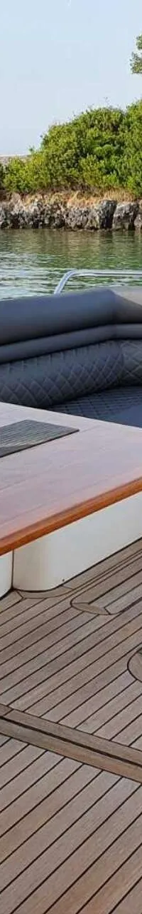 Een jachtdek met een houten tafel, donkere banken, een plantje en een Griekse vlag, met water en groen aan de horizon.