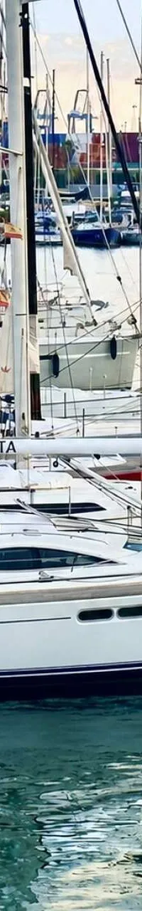 Een witte zeilboot met blauwe strepen en de naam "SPARTA" ligt aangemeerd aan een steiger, met andere boten en kranen op de…