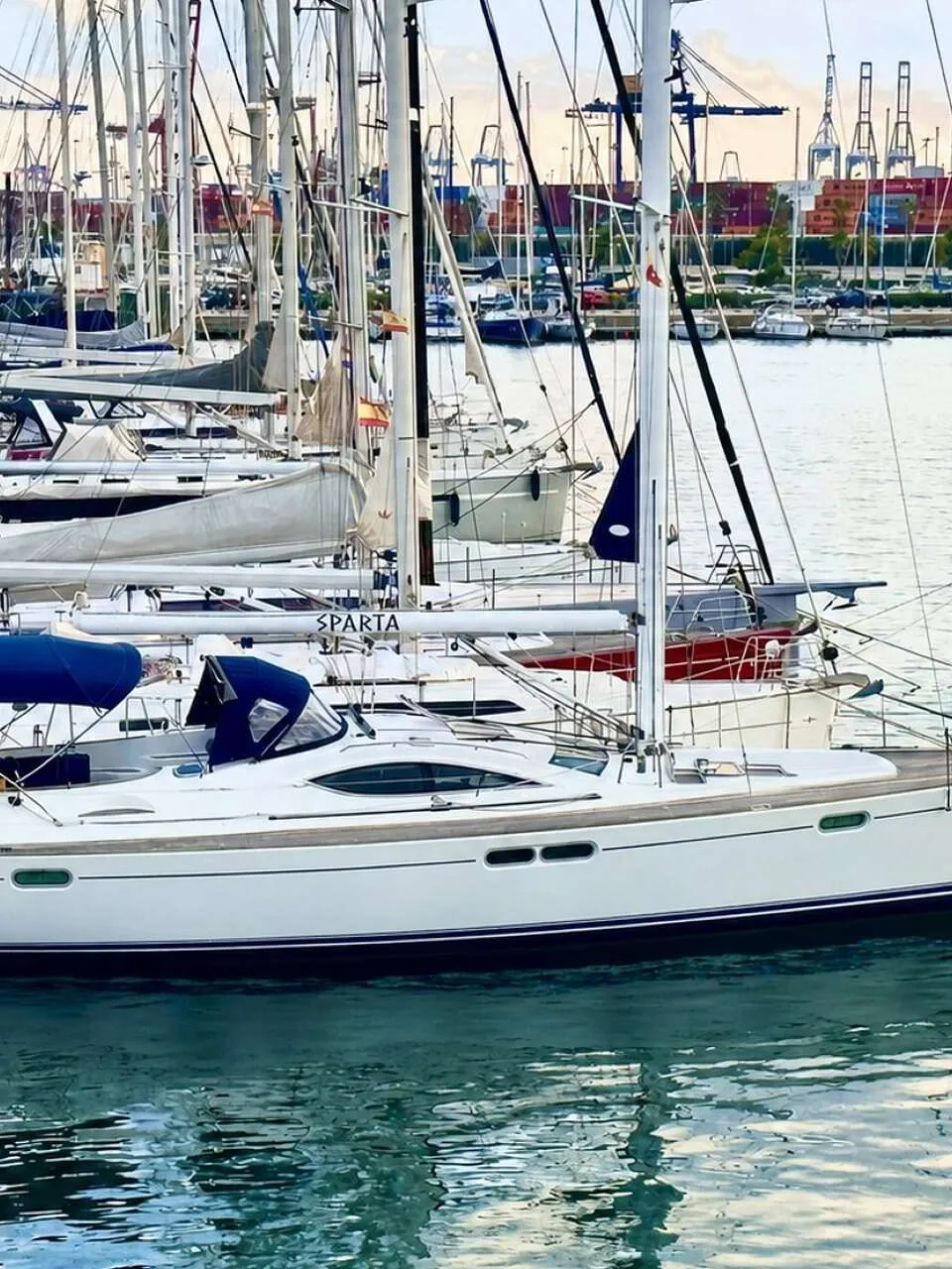 Een witte zeilboot met blauwe strepen en de naam "SPARTA" ligt aangemeerd aan een steiger, met andere boten en kranen op de…