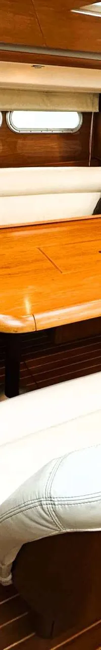 De foto toont het interieur van een boot met een houten vloer, een U-vormige witte bank met bruine kussens, een houten…
