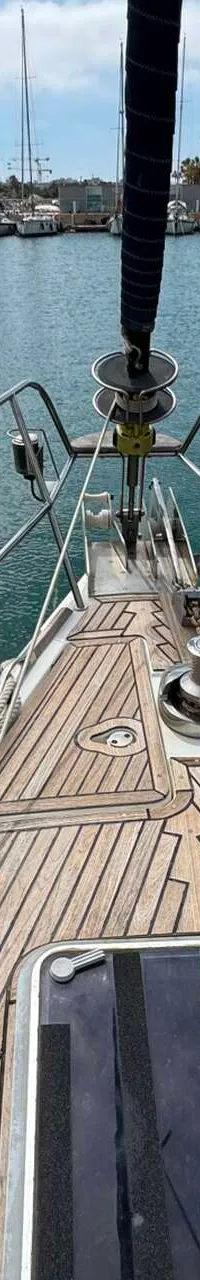 De boeg van een zeilboot met teakhouten dek, rvs reling, ankerlier en een zwart afgedekte mast is zichtbaar.