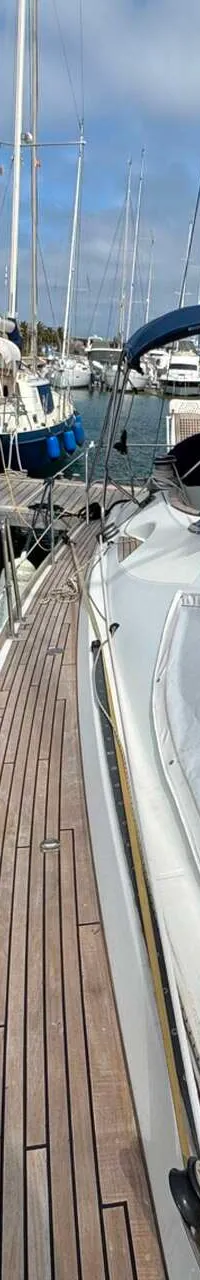 Een witte zeilboot met een blauwe buiskap ligt afgemeerd in een jachthaven, met een houten dek en andere boten op de…