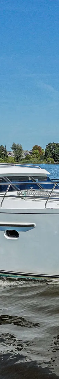 Een wit motorjacht met de naam "Harmony" vaart op een waterweg met bebouwing en bomen op de achtergrond onder een blauwe…