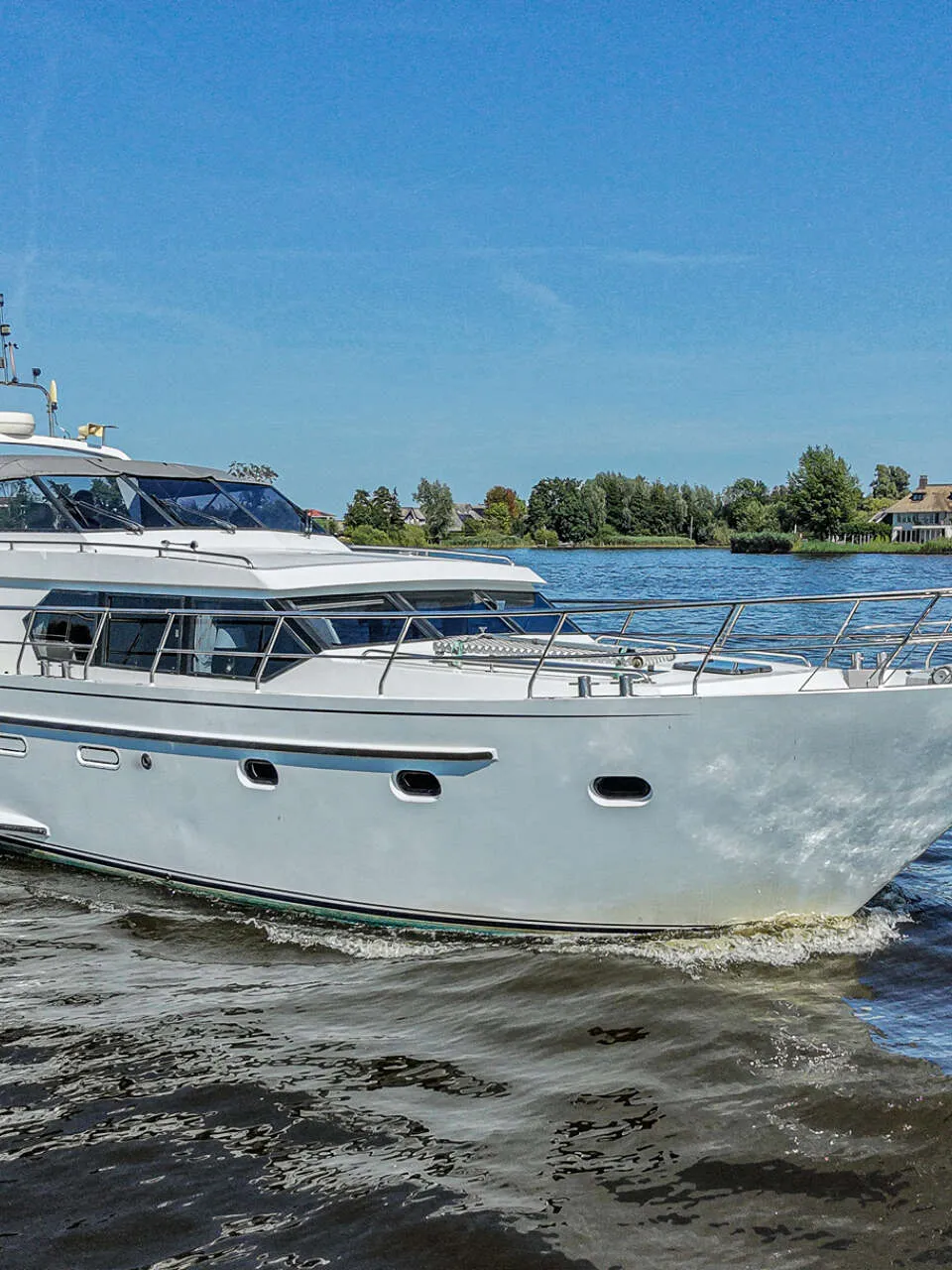 Een wit motorjacht met de naam "Harmony" vaart op een waterweg met bebouwing en bomen op de achtergrond onder een blauwe…