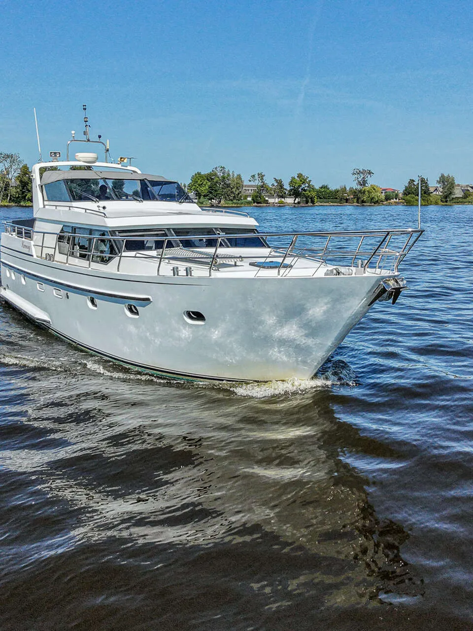 Een witte motorjacht vaart op het water met een blauwe lucht en groene oevers op de achtergrond.