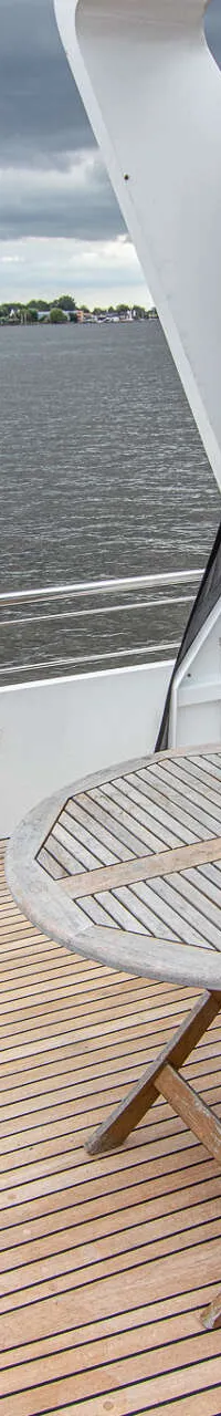 Een teakhouten achterdek met een ronde houten tafel, een blauwe bank en een open deur naar de kajuit, met water op de…
