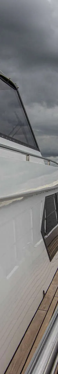 Een witte boot met een houten dek en reling vaart op water onder een bewolkte lucht, met land in de verte.