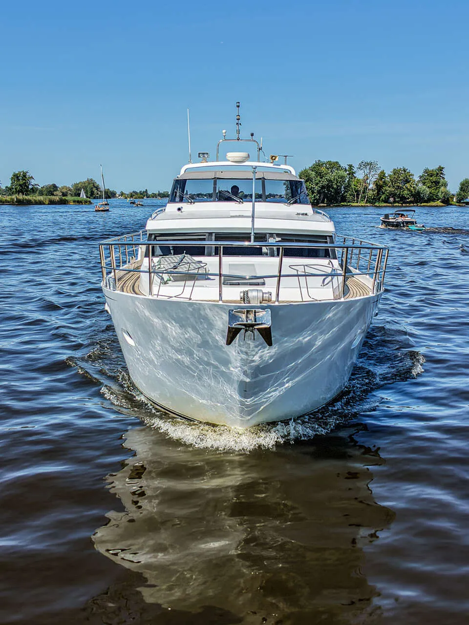 Een wit motorjacht vaart op een waterweg met bomen langs de oevers onder een helderblauwe lucht.