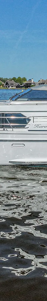 Een witte motorjacht met de naam "Harmony" vaart op een waterweg met beboste oevers onder een blauwe lucht.