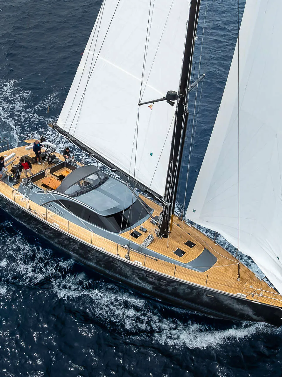 Een zeilboot met witte zeilen en een donkere romp vaart op open water, met een vlag aan de achtersteven.