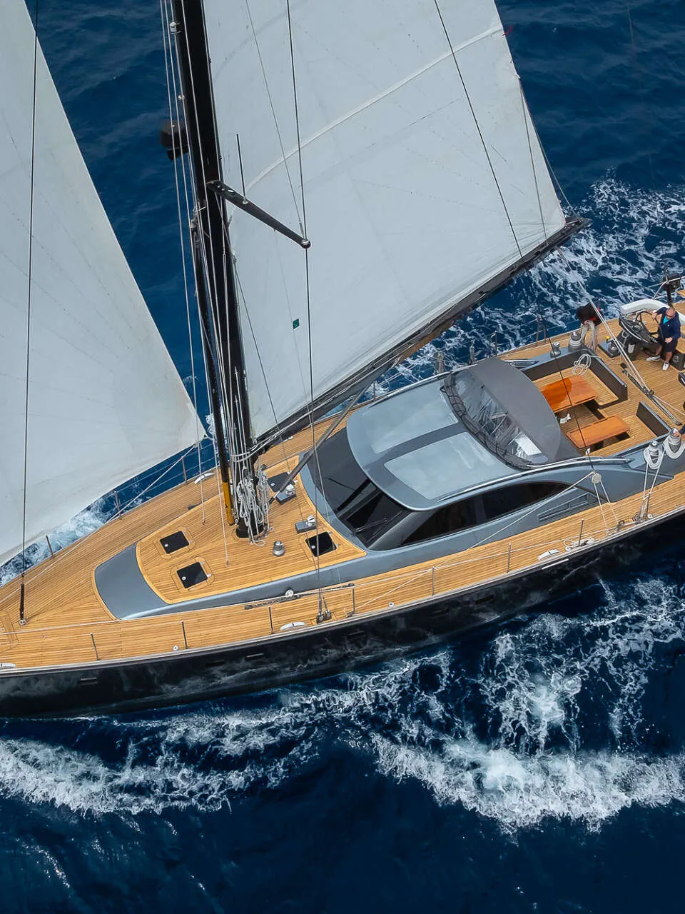 Een zeilboot met twee witte zeilen en een teakhouten dek vaart op open water met vier personen aan boord.