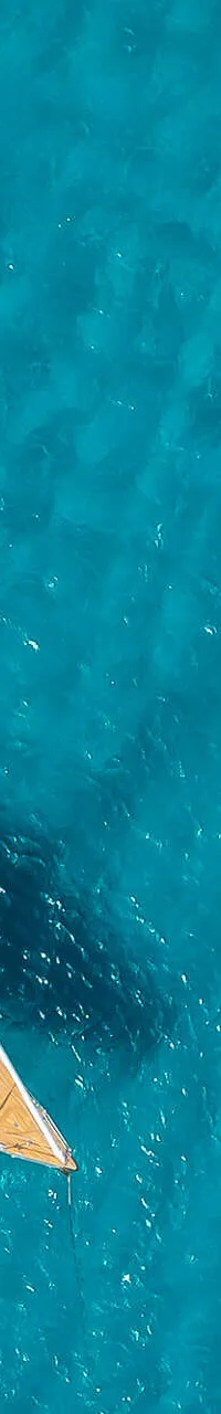 Een dronefoto van een boot met een houten dek, liggend in helderblauw water met donkere plekken.