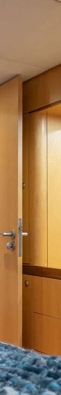 Een interieur van een boot met een bed met blauwe deken, een spiegel, houten kasten en een badkamer met wastafel.