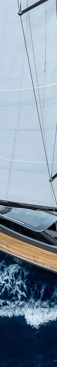 Een zeilboot met twee witte zeilen en een houten dek vaart op blauw water, met een vlag aan de achtersteven.