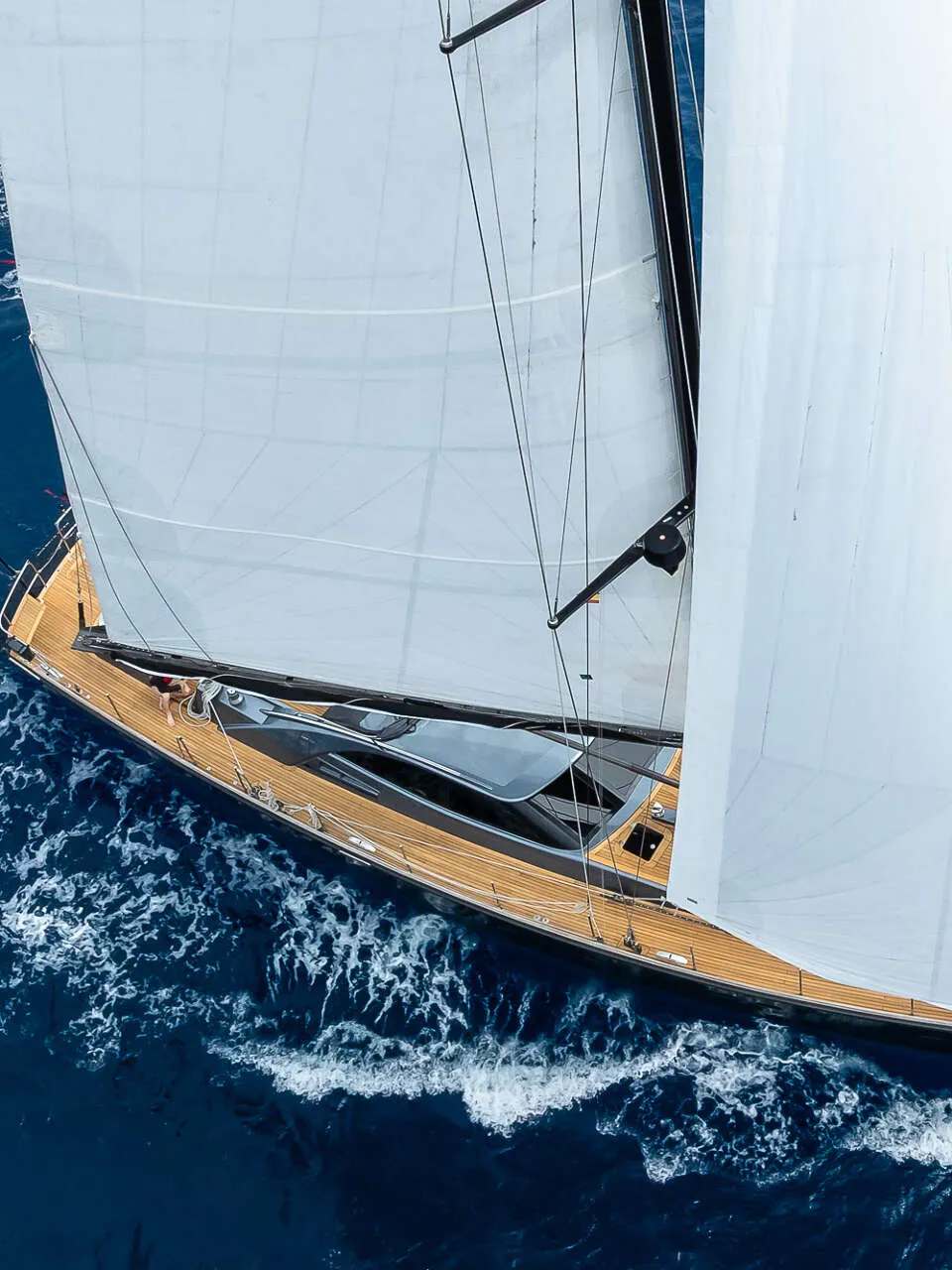 Een zeilboot met twee witte zeilen en een houten dek vaart op blauw water, met een vlag aan de achtersteven.