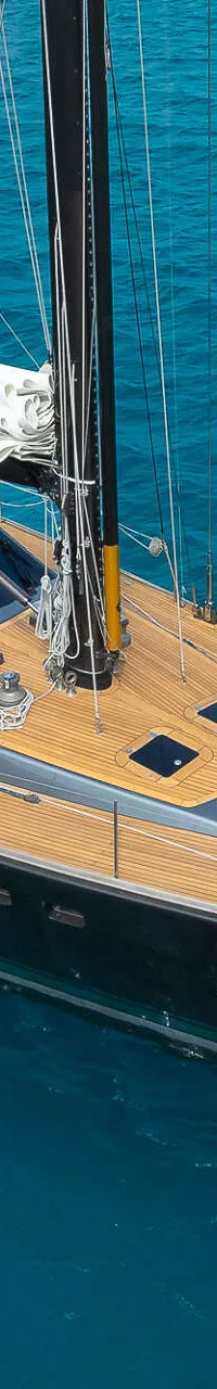 Een zeiljacht met een donkere romp en een teakhouten dek ligt voor anker in helderblauw water.