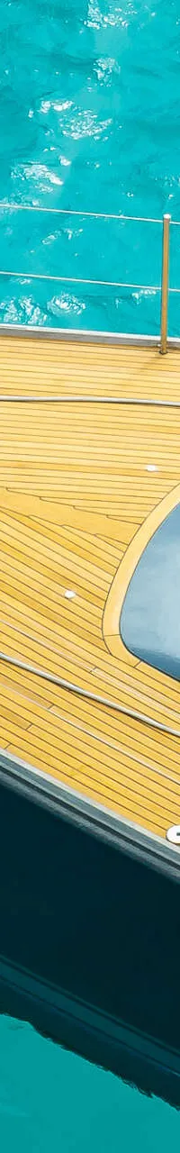 Een bovenaanzicht van een zeiljacht met een donkere romp en een houten dek, liggend in helder turquoise water.