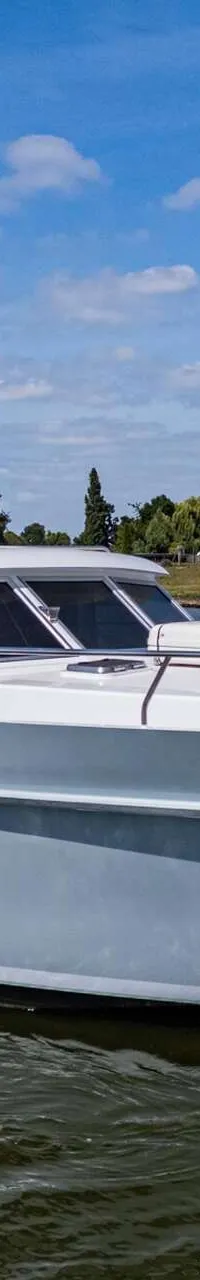 Een witte en lichtgrijze motorboot vaart op een waterweg met een groene oever en blauwe lucht op de achtergrond.
